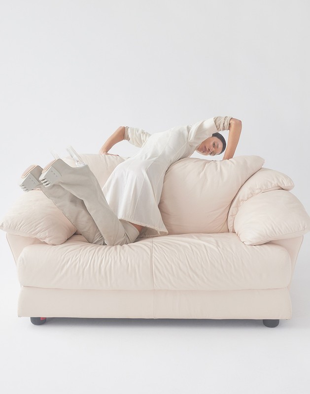 A woman in a white dress poses on a light-pink couch in a studio environment, showcasing a unique and artistic fashion statement.