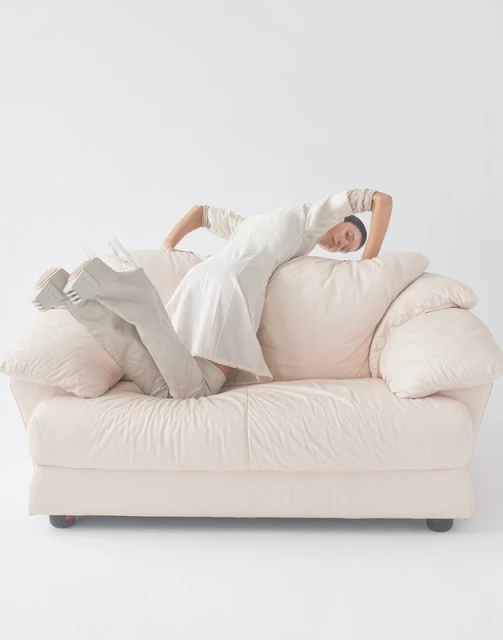A woman in a white dress poses on a light-pink couch in a studio environment, showcasing a unique and artistic fashion statement.