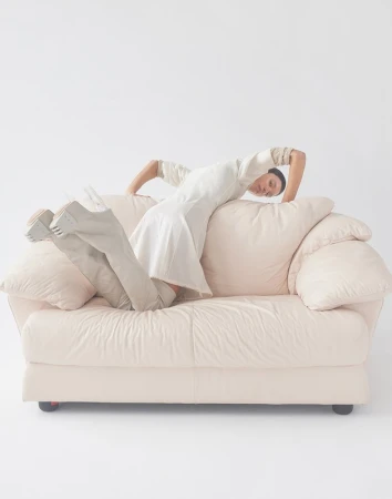A woman in a white dress poses on a light-pink couch in a studio environment, showcasing a unique and artistic fashion statement.