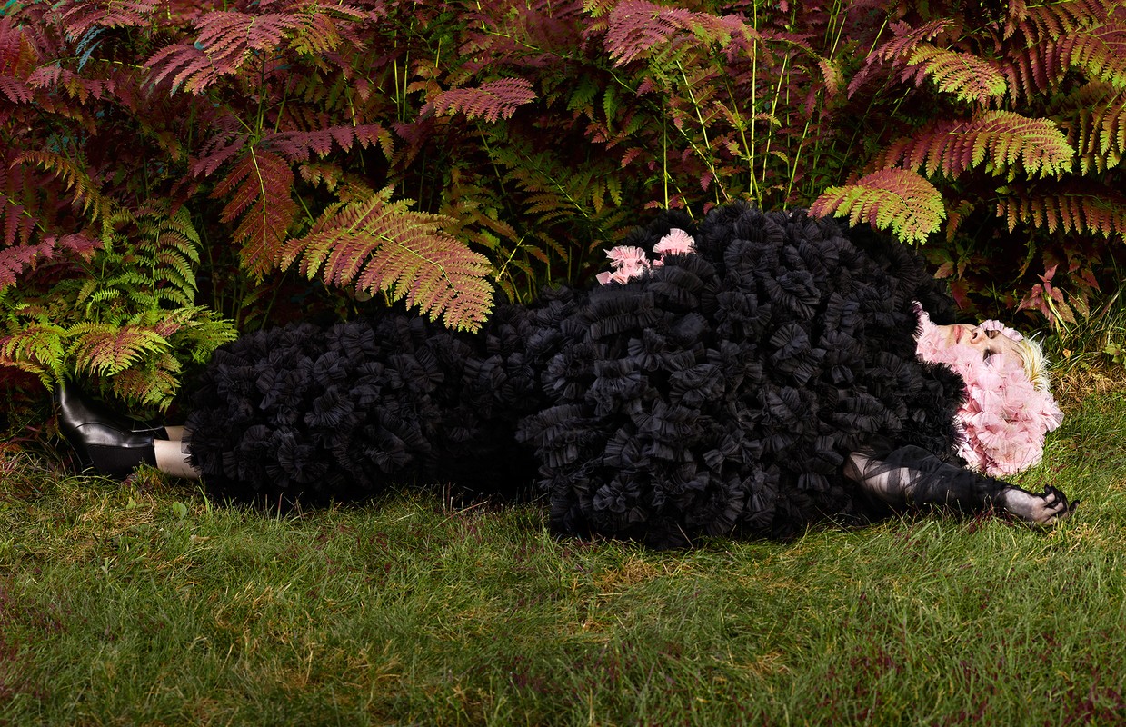 Woman in ruffled black dress lies on grass in a fashionable pose, surrounded by red and green ferns, creating a striking contrast.