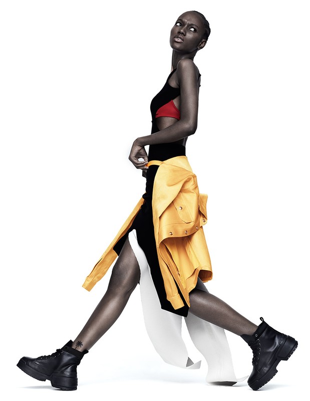 Model in black dress with yellow jacket tied around waist and black boots walking against a white background