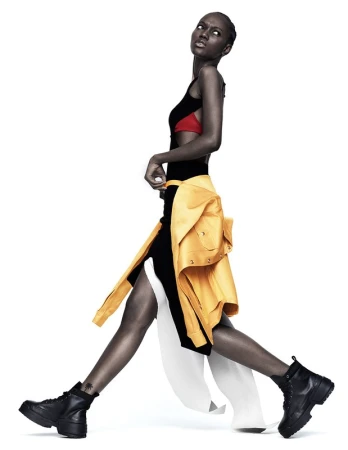 Model in black dress with yellow jacket tied around waist and black boots walking against a white background