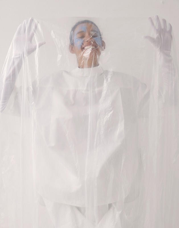 Person with painted face smiles, wearing a white top and gloves, and posed behind a clear plastic sheet, in a bright studio.