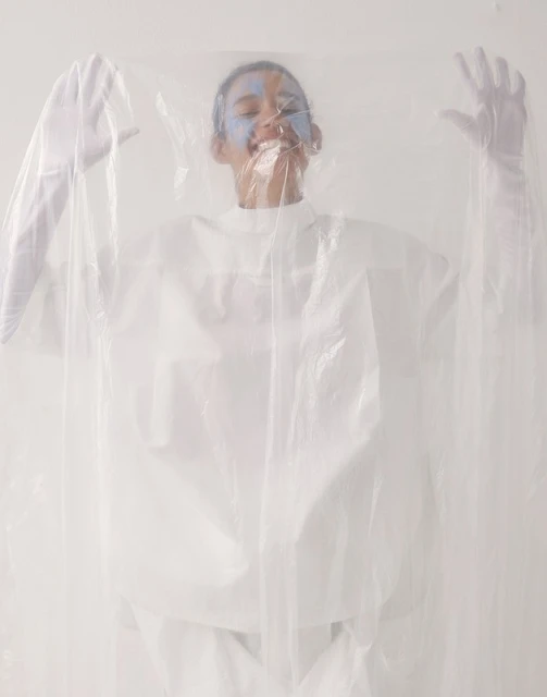 Person with painted face smiles, wearing a white top and gloves, and posed behind a clear plastic sheet, in a bright studio.