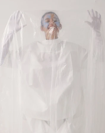 Person with painted face smiles, wearing a white top and gloves, and posed behind a clear plastic sheet, in a bright studio.