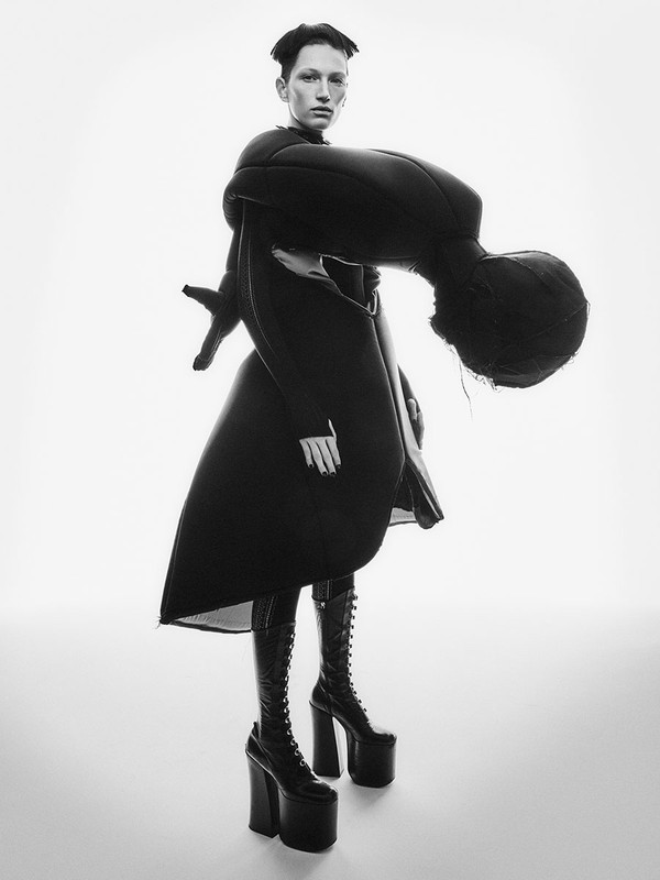 Fashion photo of a woman in black sculptural avant-garde coat and high platform boots, set against a bright white backdrop.