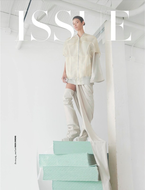 Fashion editorial shot of a person in cream-colored apparel posing on a teal structure in an all-white indoor photo studio.