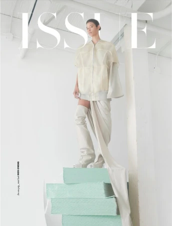 Fashion editorial shot of a person in cream-colored apparel posing on a teal structure in an all-white indoor photo studio.