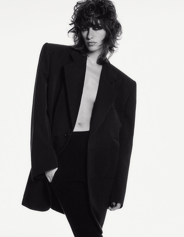 Black and white fashion shot: adult woman in an oversized black blazer and pants with wavy hair, posing in a photo studio with white backdrop.