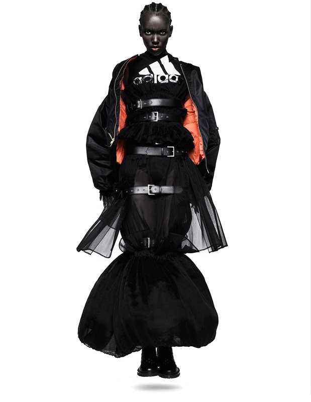 Fashion shot of a Black female model wearing an Adidas logo shirt and a black avant-garde dress with multiple belts, against a white backdrop.