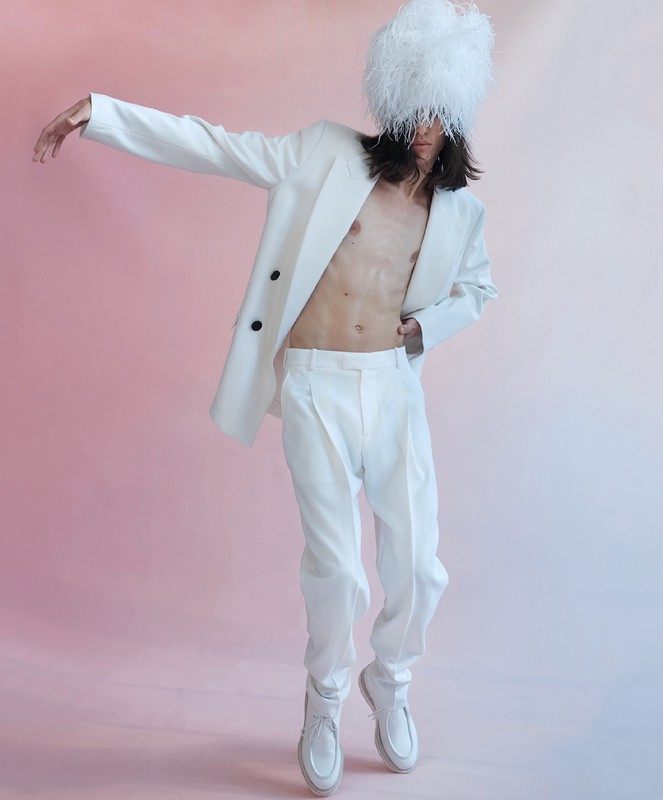 Adult male model poses in a white suit with a feathered hat against a pink backdrop in a fashion editorial shot. Studio lighting.