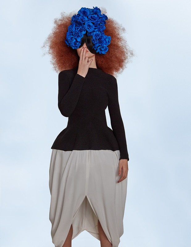 Fashionable person in black top and white tulip skirt wearing a striking blue flower headpiece against a light blue backdrop.