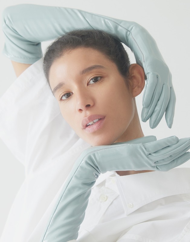 A person with light skin poses in front of a white background wearing long, pale-blue leather gloves and a white button-down shirt.