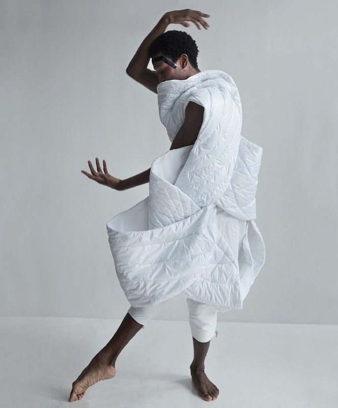 A person poses dynamically wearing a white quilted textile, barefoot, on a white seamless backdrop in a high-key fashion shot.