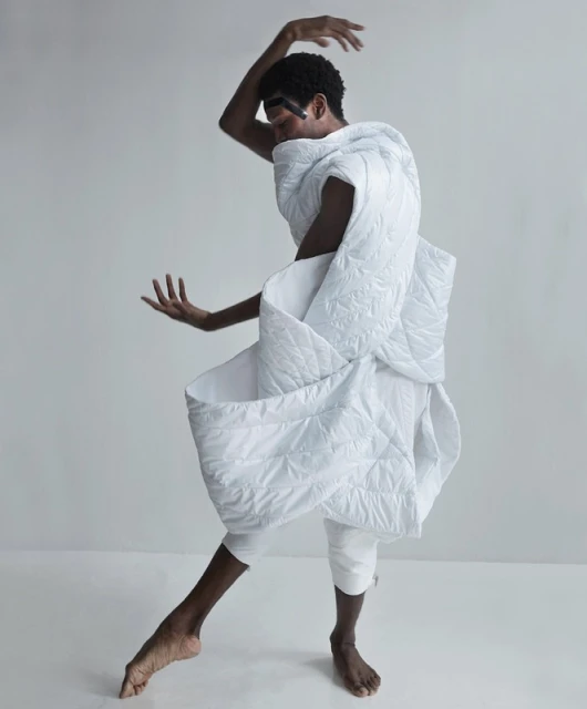 A person poses dynamically wearing a white quilted textile, barefoot, on a white seamless backdrop in a high-key fashion shot.
