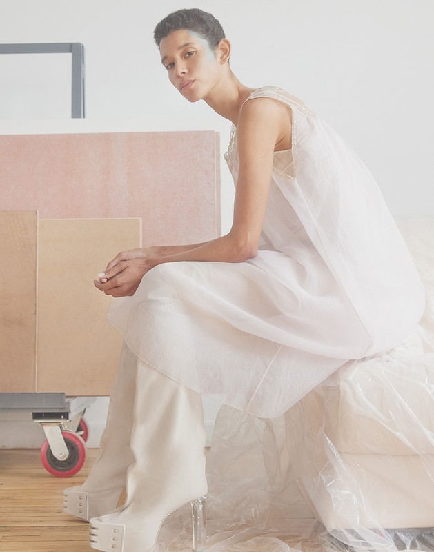 Elegant woman in sheer white dress and platform boots posing in a bright, minimalist studio setting surrounded by art supplies.