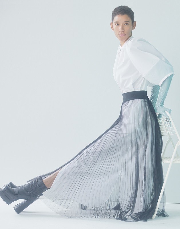 Fashion shoot of a person posing in a white blouse, a long pleated skirt and platform boots, indoors with a bright, neutral background.