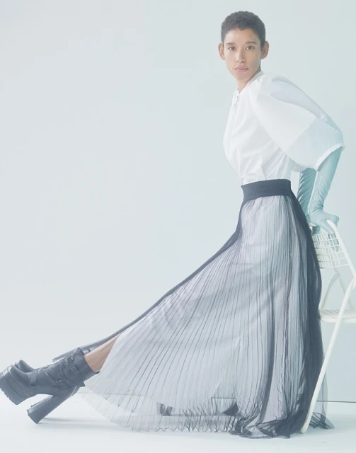 Fashion shoot of a person posing in a white blouse, a long pleated skirt and platform boots, indoors with a bright, neutral background.