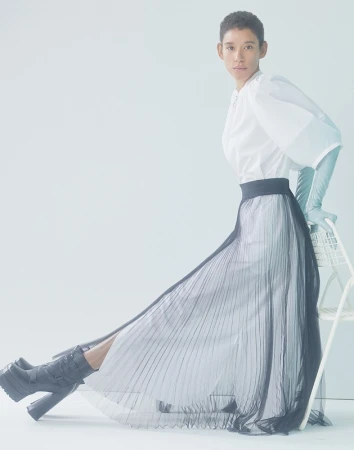 Fashion shoot of a person posing in a white blouse, a long pleated skirt and platform boots, indoors with a bright, neutral background.