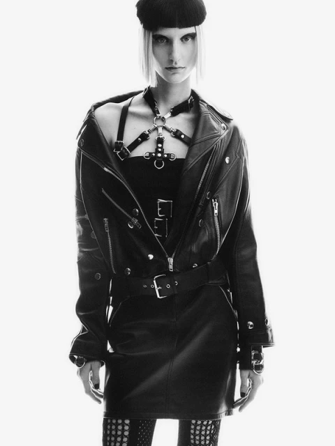 A person with fair skin and short, light-colored hair, wearing a black beret, leather jacket, harness, and skirt, poses in a studio.