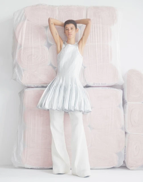 Woman poses in a halter-top dress and pants, leaning against stacks of fabric rolls in a bright studio setting, fashion editorial.