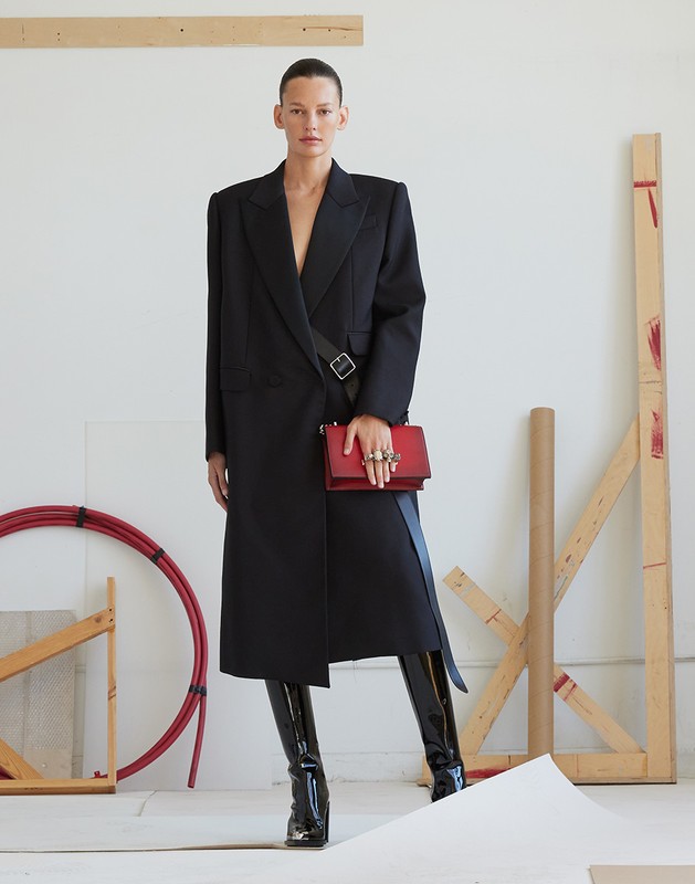 Fashion model wearing a long black coat, high-heeled boots, and red purse in a studio with red tubing and construction materials.
