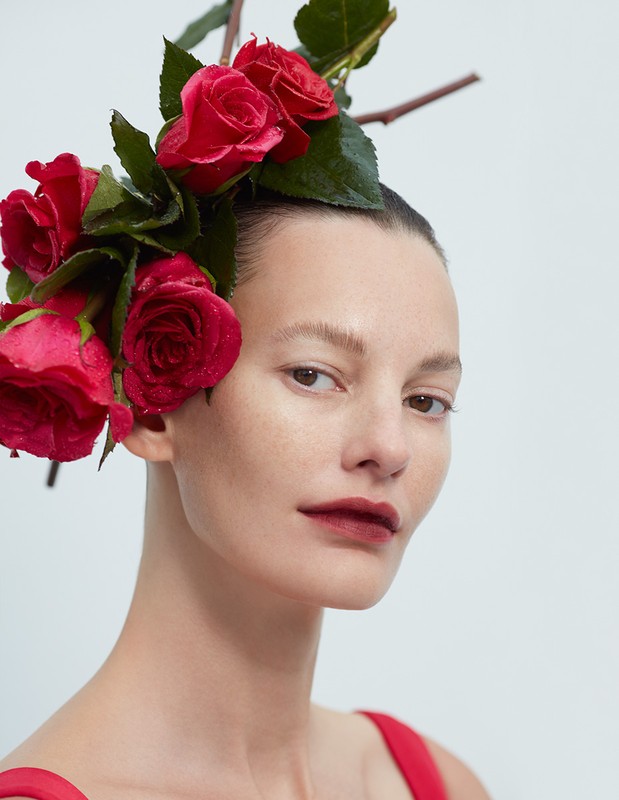 Close-up portrait of a woman with fresh red roses styled in her hair, shot in a bright indoor setting against a plain white backdrop.