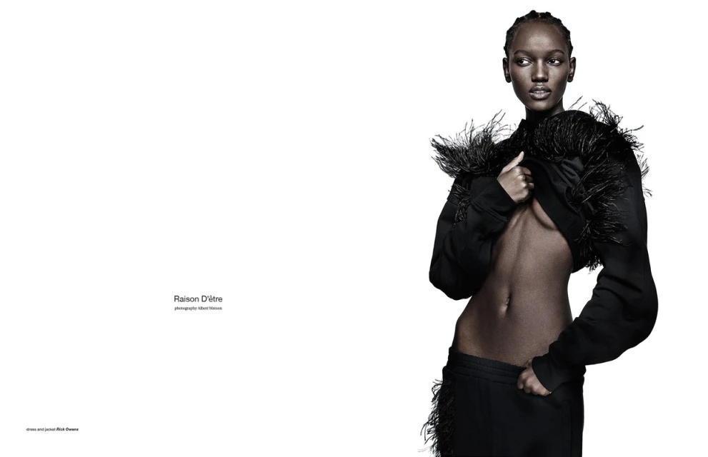 Striking fashion shot of a slim adult female with dark skin in a black jacket and pants with feather accents against a stark white background.