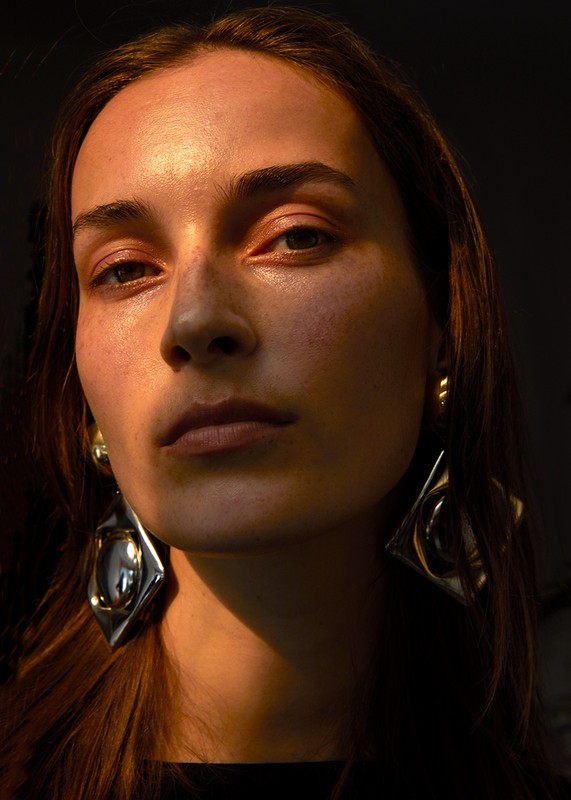 Close-up portrait of a young adult female with warm skin tones, dark hair, and large silver earrings against a dark background.