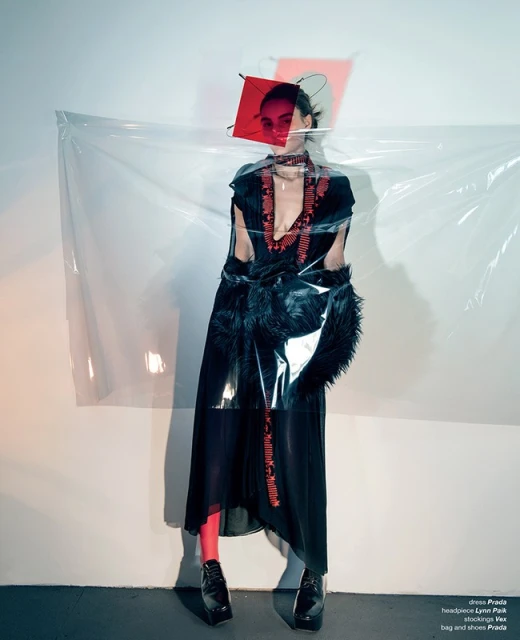 Woman in black dress and geometric headpiece poses in studio. Red tights, black platform shoes. Fashion photography in a minimalist setting.