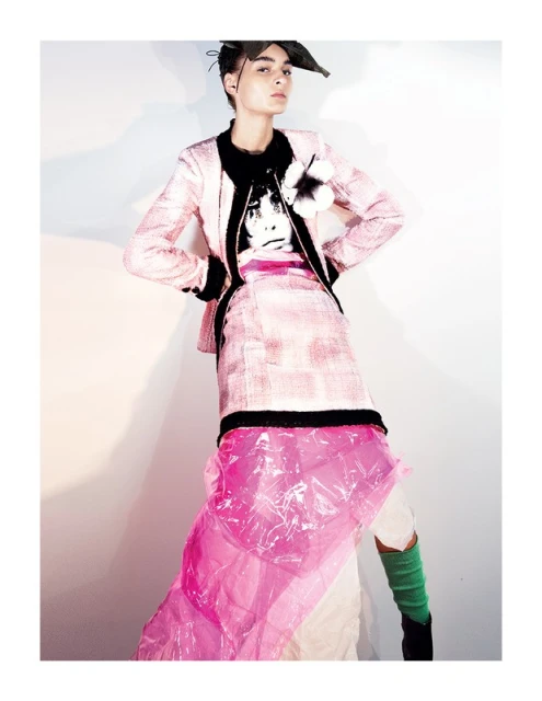 Woman poses in a pink tweed suit with a black and white graphic shirt. She is indoors against a white wall with pink plastic accents.