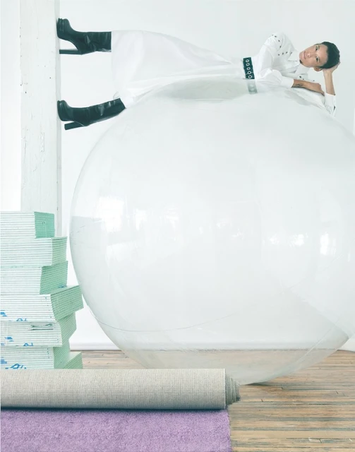 Woman in a white dress and black boots poses on a large transparent ball. A stack of boards and rolled rugs rest on a wooden floor.
