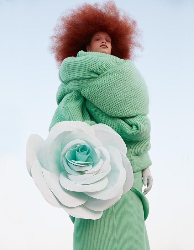Woman with a large afro in a mint green outfit with oversized knitted layers poses with a large white rose against a light blue sky.