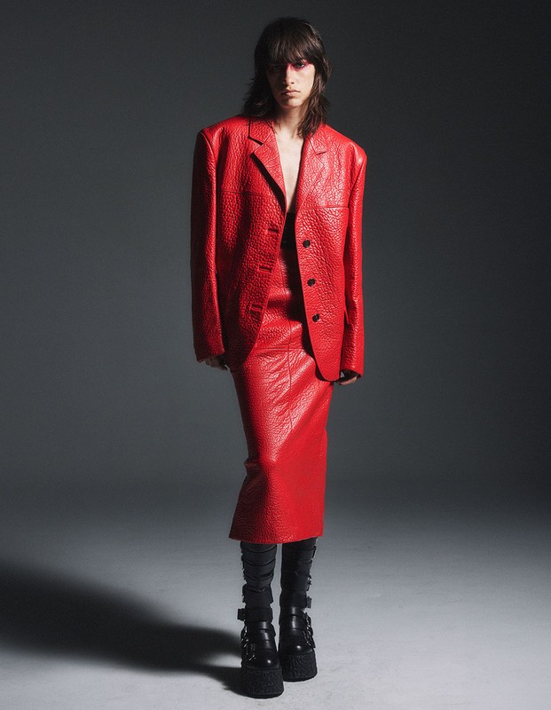 An adult female poses in a textured red suit with a blazer, pencil skirt, and black platform boots against a gradient grey backdrop.