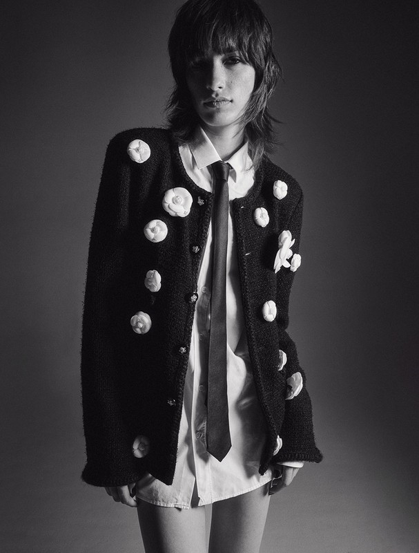 Model with textured dark hair wearing a black floral-embellished jacket, white shirt, and dark tie in a studio portrait.