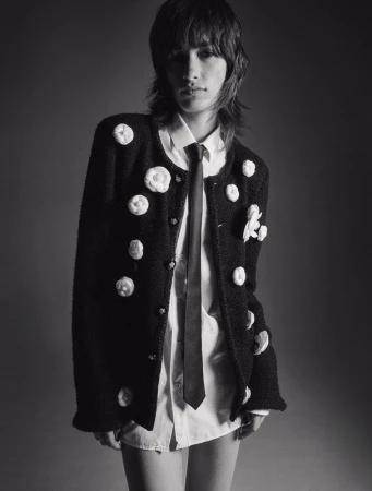 Model with textured dark hair wearing a black floral-embellished jacket, white shirt, and dark tie in a studio portrait.