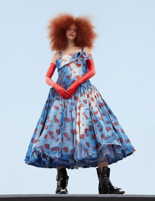 Fashionable woman with red hair, wearing a blue and red dress and black boots, posing against a light blue sky, creating a striking contrast.