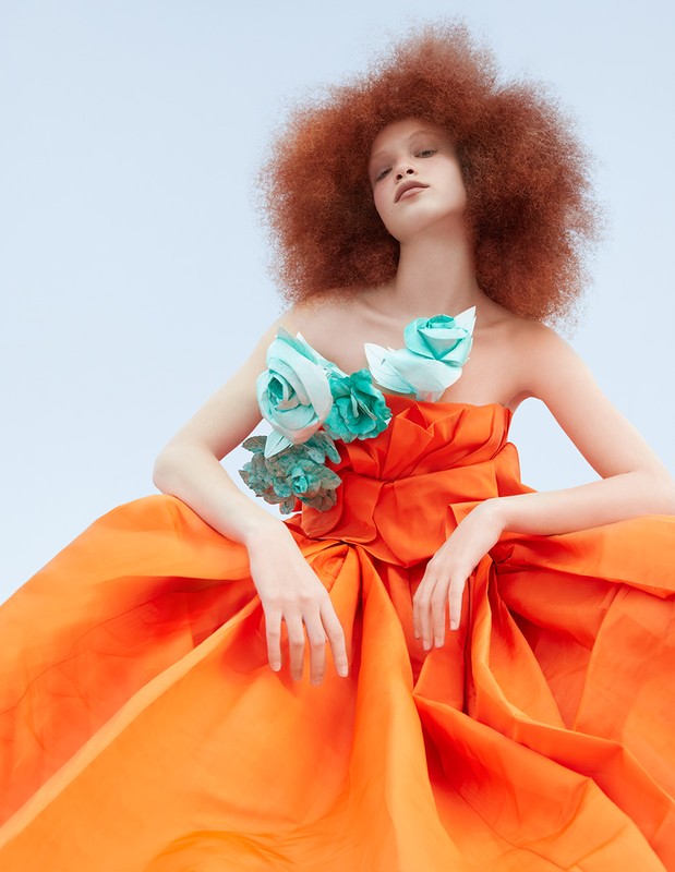 A striking fashion portrait of a woman with an afro hairstyle, wearing an orange couture gown adorned with light blue floral embellishments.