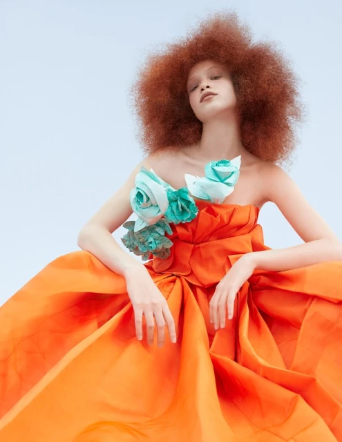 A striking fashion portrait of a woman with an afro hairstyle, wearing an orange couture gown adorned with light blue floral embellishments.