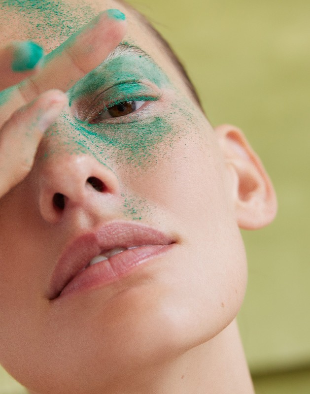 Close-up of a woman's face covered in green powder, her hand near her eye, against a yellow-green background. Beauty and cosmetic concept.