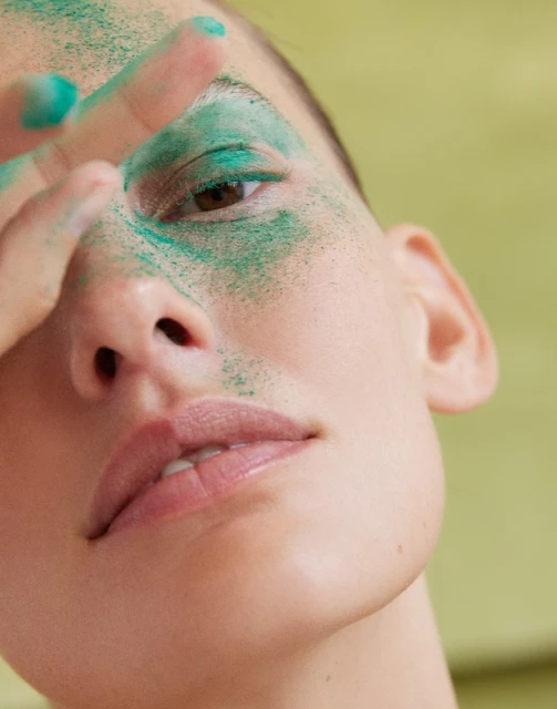 Close-up of a woman's face covered in green powder, her hand near her eye, against a yellow-green background. Beauty and cosmetic concept.