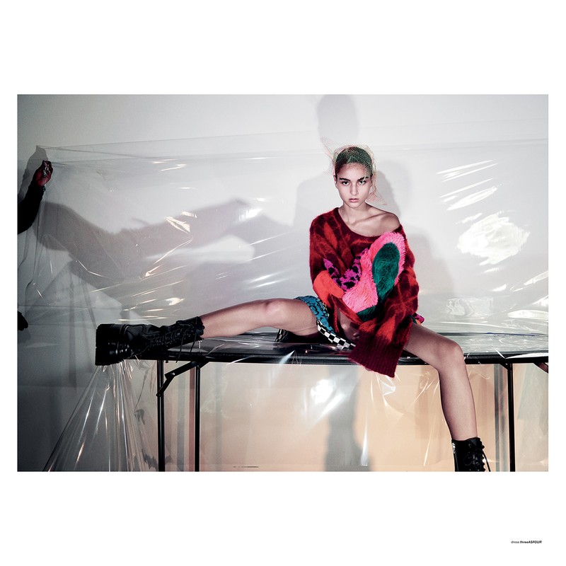 A young adult woman sits on a metal table wearing a red patterned sweater and black boots in front of plastic sheeting indoors.
