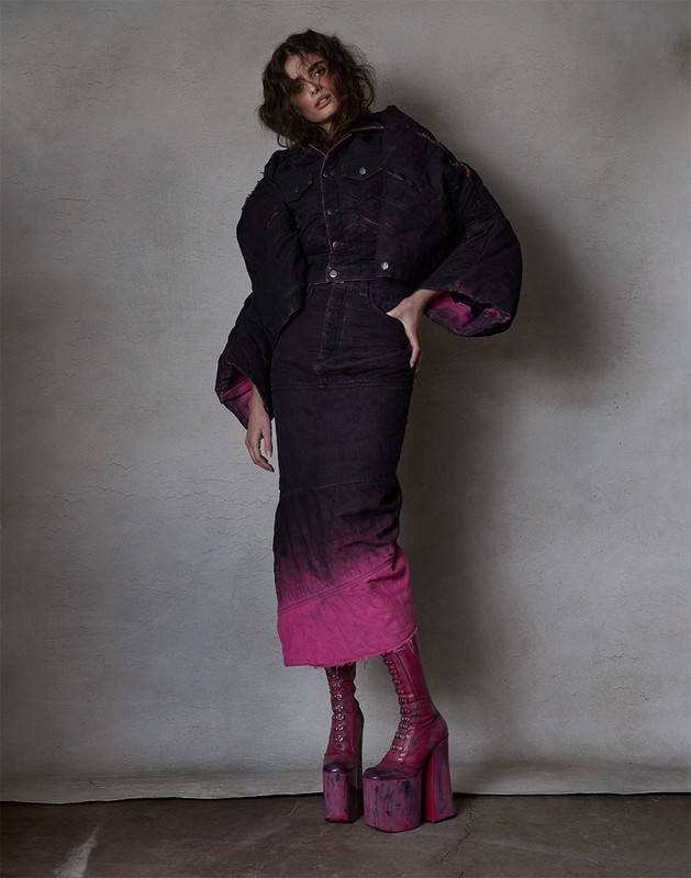 Adult female fashion model posing in a denim jacket and skirt, accessorized with platform boots, against a neutral backdrop in a studio.