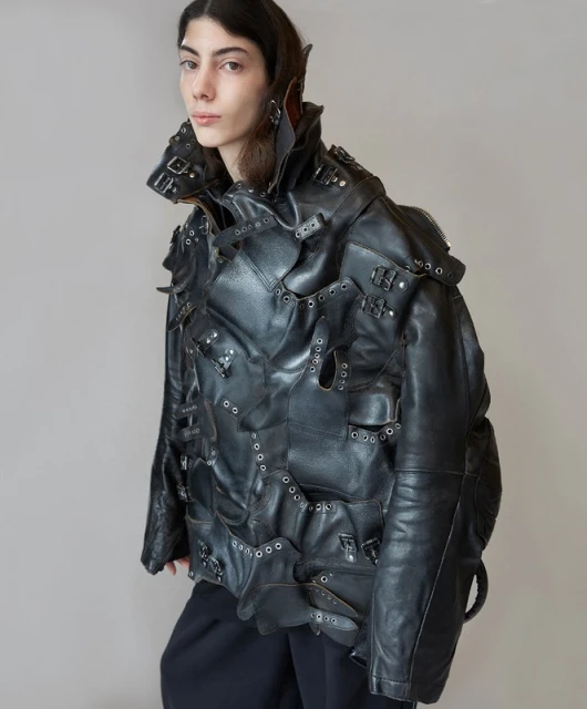 An adult female model poses indoors wearing a black leather jacket with buckles and eyelets, and dark pants, against a neutral backdrop.