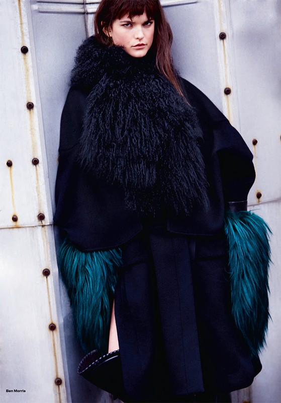 Fashion editorial shot of a woman with dark hair wearing a black coat with fur trim and green cuffs, leaning against a metal wall.