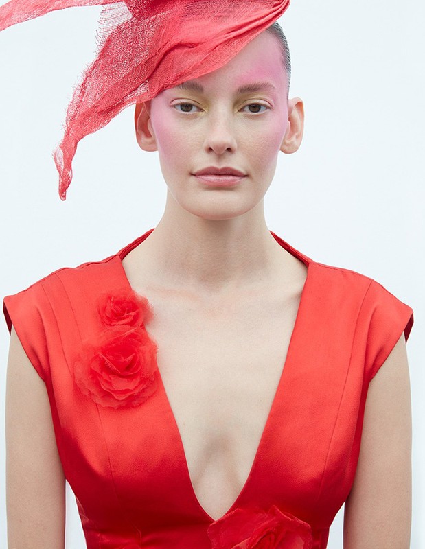 Close-up of woman in red dress with rose detail and matching red hat. Elegant fashion portrait against a clean white backdrop.
