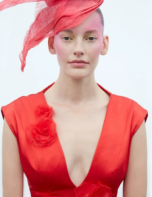 Close-up of woman in red dress with rose detail and matching red hat. Elegant fashion portrait against a clean white backdrop.