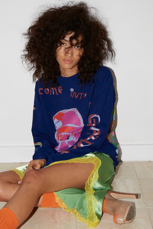 Woman with big curly hair sits on floor in blue sweater and green skirt, wearing orange socks and tan heels against white backdrop.