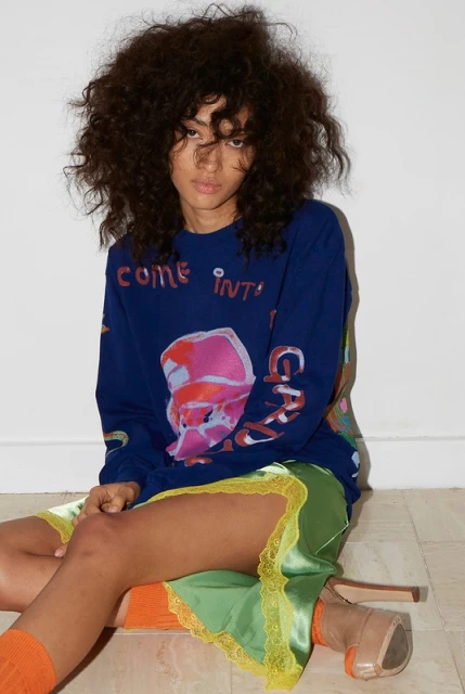 Woman with big curly hair sits on floor in blue sweater and green skirt, wearing orange socks and tan heels against white backdrop.