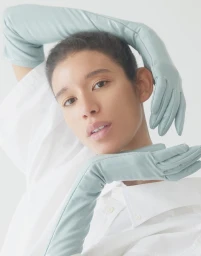 Model wearing long pale green leather gloves and a white collared shirt against a white seamless background.